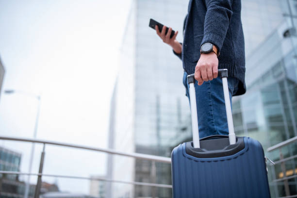 A businessman stands with his luggage and a phone in his hand.